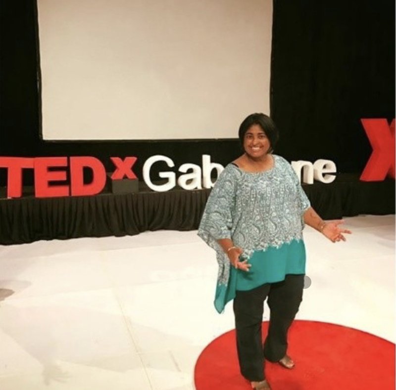 Jillian M Sigamoney on the TEDx Gaborone stage, smiling with her arms open in front of the TEDx sign.