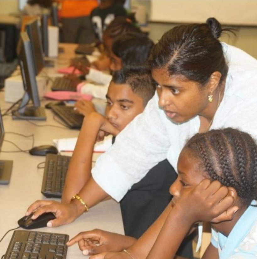 Jillian M Sigamoney leaning in at a computer to guide three students through their work.